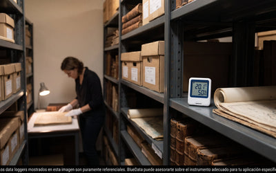 Data logger de temperatura y humedad sobre una repisa del depósito de un archivo histórico, junto a cajas de conservación y libros antiguos.
